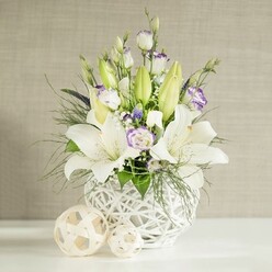 A serene, sophisticated floral display is captured as though ready to grace a wedding table in a Golders Green townhouse. At its heart stands a unique rounded white vase, its open-weave, basket-like design giving a light, airy feel, as if the flowers are suspended in a gentle breeze. From this sculptural base, tall white lilies stretch upward, some fully open with elegant trumpet-shaped blooms and visible stamens, others still held in elongated green buds that add freshness and height. Twining between the lilies are softly layered lisianthus in white and cream, their petals kissed with subtle lavender tones that look almost hand-painted. A scattering of tiny white blossoms, like gypsophila, and the occasional darker purple flower tucked deep within the arrangement lend depth without disturbing the tranquil palette. Lush foliage in varying textures - from fine, feathery fronds to broader, velvety leaves - spills casually over the rim, echoing the relaxed greenery of nearby Hampstead Heath while still feeling curated and intentional. The flowers sit on a spotless white surface, where two pale woven spheres in different sizes rest, their rattan-like texture complementing the vase and tying the composition together. The background is a soft, linen-textured wall in light beige, its horizontal pattern barely visible yet adding quiet sophistication as daylight glows across it. Everything about the image suggests gentle celebration: a table set for a wedding lunch at a venue off Finchley Road, a vow renewal at home, or a thoughtful centrepiece delivered locally by Flowers Golders Green for guests to gather around and share stories late into the evening.