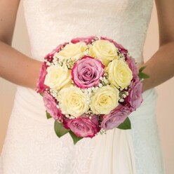 Step into the heart of Golders Green with this elegant wedding scene: a bride stands with her bouquet cradled lovingly at her waist, the soft light reflecting off roses arranged by skilled local hands. The bouquet draws the gaze-a vibrant, rounded assembly of soft, creamy yellow roses nestled with vivid fuchsia and pink blooms tinged in lilac and mauve, their petals almost glowing against wisps of snow-white baby's breath. Each element is placed with expert precision, capturing the essence of a love story told along the neighbourhood's tree-lined streets. The deep green leaves peeking out mirror the tranquil greenery of Golders Hill Park, offering balance and grounding to the design. The bride's sleeveless arms frame the bouquet, her white lace wedding dress detailed with delicate floral patterns reminiscent of timeless Golders Green bridal style. Natural daylight bathes the flowers and fabric, highlighting freshness and the intricacies of both nature and couture. This image goes beyond simple beauty-it is a celebration of community and craftsmanship, perfect for those dreaming of bespoke wedding florals that capture personal memories and local flair, ideal for wedding inspiration or sharing meaningful moments crafted by Golders Green's own floral experts.