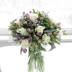 In this image, a graceful, rounded bouquet of white roses and lavender alstroemeria rests in a simple, clear glass cylinder vase, bathed in soft natural light that could be streaming through a Golders Green townhouse window. The white roses are the quiet stars of the design: fully formed blossoms with gently unfurling petals that look almost like folded silk, suggesting tenderness, respect, and a timeless sense of love. Threaded between them are numerous stems of alstroemeria, their buds and opening flowers painted in gentle shades of lavender and smoky purple, with fine streaks of deeper colour along the petals that add nuance and depth without overpowering the arrangement. Tiny clusters of baby's breath float among the main flowers, their minute white florets forming airy, cloud-like layers that catch the light and impart an almost ethereal quality. Surrounding everything is a frame of lush green foliage, from broader, darker leaves that anchor the design to slender stems that lend movement and a natural garden feel, as though the bouquet has been freshly arranged after a stroll along nearby Hampstead Heath. The stems inside the vase are cleanly cut and aligned, clearly visible through the water, showing the florist's meticulous attention to detail. The overall mood is one of stillness and hope, with the muted whites and purples conveying both comfort and celebration. It looks like a bouquet chosen for a meaningful occasion in Golders Green-perhaps to welcome a new baby, mark a thoughtful anniversary, or bring gentle solace after loss-its understated elegance reflecting the quiet care of a local floral expert.