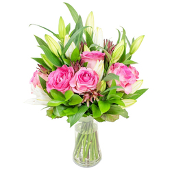 This vivid arrangement features fully opened deep pink roses and poised white lilies set into a clear glass vase, sculpted into a rounded, generous shape and shown against a pristine white field that highlights every petal and leaf. The roses dominate with layered, velvety petals in luscious tones of fuchsia and soft blush, their form both romantic and exuberant. Nestled among them, white lily buds present a sequence of stages-some closed with neat, tapered tips, others just beginning to unfurl their delicate trumpets-introducing a quiet promise of evolving fragrance and form. Lush green foliage, from broad glossy leaves to slender, lighter-green sprigs, provides volume and a cooling verdant backdrop; the foliage's varied textures deepen the composition and frame the central blooms. Scattered through the greens are darker reddish-brown accents-small buds and filler stems-that give weight and a subtle autumnal note to the palette. The transparent vase, slightly flared at the rim and narrowing toward the base, reveals crisp, trimmed stems descending into clear water, a small, honest detail that underscores the bouquet's freshness and care. The photograph evokes a calm, celebratory mood suitable for birthdays, Mother's Day, or simply brightening a home near the shops around Golders Green Road. The arrangement reads as the work of a practiced hand-balanced, generous and deliberately joyful-inviting the viewer to imagine the soft scent of roses mixing with the clean, floral breath of lilies in a quiet London morning.