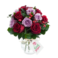 A richly coloured floral arrangement, densely rounded and meticulously balanced, combining deep crimson and burgundy roses with delicate mauve and dusty-lilac roses to create a sophisticated two-tone harmony. Each rose is fully cupped and freshly opened, the petals exhibiting that soft satin sheen that invites touch, while the dark green foliage - likely Ruscus or similar evergreen leaves - sits between blooms to accentuate the colour contrast and add a cool, leafy fragrance. Scattered through the composition are small clusters of red-orange berries or rose hips, their glossy skins and compact forms adding a playful, autumnal texture that offsets the roses' velvety softness. The bouquet is held in a rounded mercury-effect glass vase with a slightly bulbous base and tapered neck; its antiqued silver surface mirrors flecks of the roses' tones and is finished with a simple natural twine bow. A rectangular white tag dangles from the neck, showing a small hand-drawn rose motif and space for a handwritten message rather than a commercial label, which keeps the sentiment intimate. Set against a pristine white background, the arrangement reads as both luxurious and homey - ideal for bringing to a celebration in a private Hampstead sitting room or for same-day hand delivery across Golders Green when you want to say something heartfelt without words.