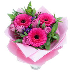 A lively trio of fuchsia Gerbera daisies takes centre stage in this vibrant bouquet, their saturated petals fanning outward to create a cheerful focal point. Nestled closely around these are softly hued lavender-pink roses, their petals curled inward like private promises, bringing tenderness and a refined softness to the composition. Interspersed among the larger blooms, clusters of small purple statice add a delicate, papery texture and a cool contrast that makes the pinks sing. The arrangement is grounded by generous green foliage - broad leaves with a faintly glossy surface that suggest salal or eucalyptus - which gives the bouquet fullness and a fresh, lightly herbal aroma.

This hand-tied bunch is presented with thoughtful finishing touches: an inner layer of pale pink, matte tissue paper cushions the stems while an outer wrap of sheer, slightly crinkled fuchsia-pink cellophane lends a gentle shimmer and reinforces the pink palette. The clear glass vase rim peeks out below, indicating the bouquet is ready to be placed on a dining table or reception desk without further fuss. The scent is soft and comforting - roses offering a classic floral perfume, gerberas a quieter green sweetness - creating an approachable fragrance profile that suits celebrations or quiet, heartfelt moments alike. As a local Golders Green arrangement, it's ideal for sending warm wishes to friends and family near the Tube station or for same-day delivery within Golders Green and neighbouring Hampstead. The overall effect is joyful, intimate, and expertly composed.