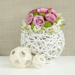 On a smooth white tabletop, a carefully composed floral centrepiece commands attention, the sort of refined design often chosen for wedding breakfasts and engagement dinners around Golders Green and nearby Hampstead Garden Suburb. The arrangement is cradled in a distinctive round vase made of white, rattan-style strands, woven loosely into an open sphere that lets light pass through the gaps, creating gentle shadows beneath. From this sculptural base rises a full, domed bouquet of roses in shades of soft pink and muted lavender, each bloom layered with velvety petals that curl slightly at the edges, some tinged with a deeper blush as though brushed with watercolour. Between the roses, bright green button mums sit like little cushions, their tiny, packed petals bringing playful texture and a clean, fresh hit of colour. At the base, clusters of dainty white filler flowers and cream-toned blossoms form a delicate fringe, giving the design a light, frothy halo as they spill just over the rim. To the left, two decorative balls made from woven, pale wicker rest on the tabletop, one larger than the other, their organic latticework echoing the vase and creating a quiet rhythm in the composition. Behind, a softly textured beige backdrop, similar to natural linen or fine burlap, is gently out of focus, offering warmth without drawing the eye away from the blooms. The soft, even lighting enhances every curve of petal and twist of twig, making this piece feel serene, romantic, and thoughtfully crafted for an intimate wedding reception or stylish home gathering in the Golders Green area.