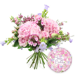 A softly lit, celebratory image shows a hand-tied bouquet and coordinating birthday balloon resting against a pure white backdrop, giving the composition a fresh, airy feel. The bouquet is generously rounded, dominated by several large, full heads of blush pink hydrangea whose innumerable petals overlap to create a plush, cloud-like effect. In and among these hydrangeas nestle pink roses and tiny spray roses; some are in full bloom, revealing elegant spirals at their centres, while others remain in tight, rounded buds that lend a sense of promise. Threads of deeper fuchsia or magenta waxflowers wind through the larger blooms, their minuscule blossoms and slender stems adding depth and contrast, rather like the small details one notices when walking through the quieter back streets of Golders Green. Lightly lifting above the main flowers, campanula stems carry small, bell-shaped blooms in shades of lavender and periwinkle blue, giving vertical rhythm and a cool note against the warmer pinks. A mixture of fresh green foliage surrounds the bouquet, providing structure and a natural, garden-inspired frame. At the base, the stems are neatly gathered and visible, highlighting the professional hand-tied technique used by local florists. To the right of the bouquet, slightly below its midpoint, sits a round, metallic "Happy Birthday" balloon whose sheen catches the light. Its surface is illustrated with a pattern of petite roses and buds in soft pink, lilac, and cream, mirroring the bouquet's palette, while the central inscription reads "Happy Birthday" in graceful purple script. The arrangement and balloon together evoke a sense of warmth and celebration, perfect for marking a birthday in Golders Green and the surrounding North West London neighbourhoods.