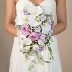 What unfolds in this evocative scene is an elegant, teardrop-shaped bridal bouquet crafted by the experienced hands of a Golders Green florist, offering a sensory symphony of gentle pastels and luminous whites set against a tranquil, neutral background. The bouquet, held close by a bride in an intricately lace-embellished, strapless gown, becomes a living poem of bridal floristry. Luminous white phalaenopsis orchids with glowing yellow throats float serenely atop layers of lush, delicate pink garden roses and tightly bunched lavender stock, their textures intermingling in a way reminiscent of a slow morning walk past the Golders Green Hippodrome. Lisianthus blooms, their white petals edged with touches of purple, nod softly amid accents of airy baby's breath, infusing the bouquet's cascade with a gentle, romantic movement. Flecks of fresh greenery, subtle yet present, peek from the floral abundance, much as glimpses of nature might be spied through the leafy lanes around Hampstead Garden Suburb. As the bouquet spills gracefully downward, it echoes both the flowing silhouette of a wedding dress and the timeless elegance so often sought for marriages in Golders Green. The whole arrangement exudes an understated, clean fragrance and a sense of tranquil celebration-an embodiment of local artistry designed for unforgettable wedding moments shared with family and friends in North West London.