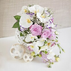 On a smooth white surface, an opulent floral centerpiece draws the eye with its serene mix of whites, blush pinks, and soft purples, much like a sophisticated wedding table set for guests in Golders Green. The arrangement is gathered into a distinctive spherical white vase, its open weave resembling fine wicker or a delicately wound ball of twine. From this airy base, arching stems of white Phalaenopsis orchids emerge, their petals wide and velvety, each bloom showing a tiny burst of golden-yellow at its center that lends an exotic, almost luminous detail. Surrounding the orchids, pastel pink and lavender roses open fully, their ruffled layers suggesting fragrance and romance, ideal for a bridal table or an anniversary dinner in a nearby restaurant. Scattered between them are smaller blossoms of Lisianthus or Eustoma, their white petals gently edged with lilac or deeper purple, giving the design a soft watercolor effect. Wisps of white baby's breath weave through the flowers like fine mist, adding volume without weight, while pale purple stock or similar trailing blooms spill slightly downward, hinting at a relaxed, organic movement. Deep and mid-green foliage grounds the palette, framing the lighter tones. To one side, two decorative woven balls in cream and white repeat the vase's texture and provide a sculptural accent on the otherwise minimalist tabletop. The background is a light, neutral beige wall, subtly textured and softly blurred, suggesting a calm interior somewhere off Finchley Road. The overall mood is tranquil yet celebratory, the kind of carefully crafted piece a Golders Green florist might create for a chic wedding, engagement party, or elegant home gathering.