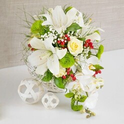 A luxurious white and ivory floral centerpiece, styled as if for an elegant Golders Green wedding breakfast, rests in a round white wicker basket on a spotless white tabletop. The arrangement is dominated by several magnificent white lilies, their broad trumpet petals curving outward and catching the light, while delicate speckling near the throats and gently arching stamens add a hint of drama. Tucked among the lilies, a single creamy yellow rose forms a romantic focal point, its spiral of petals tightly gathered yet hinting at a soft fragrance. From the right side of the display, white phalaenopsis orchids tumble in a graceful cascade, each bloom perfectly formed with a porcelain-like sheen, suggesting the fluid line of a bridal gown. Around these star flowers, fine feathery greens, glossy leaves, and playful bright green dianthus balls create a layered, tactile backdrop, evoking the lush greenery of the paths leading up towards Golders Hill Park. Tiny white baby's breath flowers lace through the design, adding a powdery softness, while scattered clusters of deep red berries bring jewel-like highlights that feel celebratory yet refined. The arrangement sits against a warm, neutral fabric background in pale beige, which gently frames the composition without stealing attention. On the left, two white decorative spheres made from interwoven strips echo the basket's texture and provide a modern, sculptural touch, reminiscent of contemporary interiors on Golders Green Road or Finchley Road. The overall mood is one of polished serenity and joyful expectation, an ideal table arrangement for anniversary dinners, intimate receptions, or stylish celebrations at home with fresh flowers delivered locally the same day.