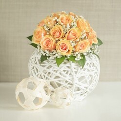 A captivating floral display shows a domed cluster of around two dozen peach and apricot roses, each one carefully placed so the blooms form a smooth, rounded silhouette that looks almost like a glowing sunrise captured in flowers. The inner petals of the roses carry a richer, deeper orange tone, while the outer petals soften into creamy peach, giving the bouquet a layered warmth that feels inviting and comforting. Between these velvety roses, fine sprays of white gypsophila-baby's breath-float like tiny clouds, their frothy texture lightening the design and adding a sense of movement, as if a gentle breeze had just brushed through. Small hints of dark green foliage hide beneath the blossoms, bringing contrast and a natural, garden-fresh feel that suggests the flowers were chosen at their best, much like a careful selection from a weekend market near Golders Hill Park. The arrangement is held in a modern spherical vase made from white woven strips, its open lattice pattern twisting and crossing to form an airy globe that feels both sculptural and delicate. Through the gaps of the weave, you catch glimpses of stems and shadow, which add an extra layer of interest. On the smooth white tabletop in front, two smaller decorative orbs, fashioned from pale, natural-looking material, repeat the same woven theme, one slightly larger than the other to create a balanced trio. Behind everything, a light beige or greige wall with a fine vertical texture serves as a calm backdrop, ensuring the warm rose tones and crisp whites remain the focus. Soft, diffused daylight illuminates the composition, evoking a sense of quiet celebration perfectly suited to an intimate dinner in a Golders Green home or as an elegant gift arrangement available from local florists for same-day delivery across the neighbourhood.