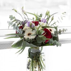 Within the sun-drenched calm of a Golders Green morning, a bouquet comes to life, showcasing the understated charm and vivid hues found in North West London's finest gardens. The bouquet's core is anchored by deep red roses, their folds luxuriously velvety, creating a passionate focal point softened by the crisp elegance of large white gerberas whose dark centers lend a touch of comfort and steadiness. Soft lilac lisianthus, some unfurled and others just at the cusp of opening, bring hints of promise and gentle movement, echoing the quiet anticipation of new beginnings. Wisps of fluffy white filler blooms fill the spaces like dewdrops clinging to grass at nearby Golders Green Park. Eucalyptus leaves arc gracefully, their silvery-green discs bringing a breath of forest-like freshness and local authenticity, while the entire composition is wrapped with dark green, broad-leaf foliage, framing the arrangement as beautifully as the tall trees lining Hoop Lane. Resting in a clear glass vase, the bouquet's stems and the fresh water below are visible in a nod to purity and craftsmanship. The aroma-a subtle interplay of rose and eucalyptus-evokes strolls past Brent Cross's bustling flower corners. Ideal for celebrations, comfort, or everyday appreciation, this arrangement provides an invitation to pause and soak in the peace and artistry emblematic of Golders Green's welcoming spirit.