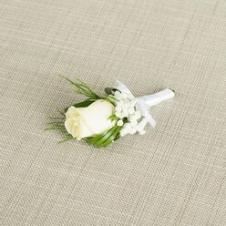 A refined wedding boutonniere is carefully placed on a neutral, light beige fabric that looks like finely woven linen, its subtle diagonal texture providing a calm, natural backdrop reminiscent of an elegant reception venue tucked off Golders Green Road. The focal point is a single ivory-white rosebud, medium-sized and perfectly formed, its petals smooth and velvety, curving tightly inwards with just the slightest suggestion of unfolding at the tip. Around the rose, delicate sprigs of white baby's breath create a halo of tiny, star-like blossoms, their airy clusters lending a cloud-soft texture and a romantic softness to the composition. Slim, pointed leaves in rich, polished green extend discreetly beyond the blooms, while a few feathery, fern-like fronds peek through, adding movement, depth, and a whisper of woodland charm, as if the foliage had been freshly gathered from a sheltered garden near Hampstead Heath. The stems of the rose and greenery are expertly bound together with a crisp white satin ribbon, its surface smooth and faintly reflective under the gentle light. The ribbon is wrapped tightly and evenly, then fashioned into a neat, modest bow positioned just beneath the rose, giving the boutonniere a finished, tailor-made look suitable for the sharp lapel of a groom's suit. Soft, diffused lighting bathes the scene, illuminating the creamy tones of the bloom, the snowy gypsophila, and the lush greens without overpowering them. The overall effect is one of classic, understated luxury and heartfelt tenderness, a small yet meaningful floral detail ideal for weddings, vow renewals, or formal celebrations across Golders Green and nearby Temple Fortune.