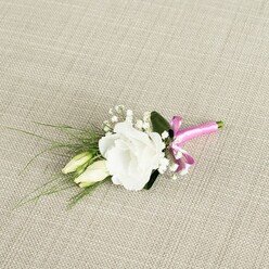 A finely crafted boutonnière lies diagonally across a pale beige, linen-like fabric, the textured weave forming a calm, neutral stage for a single, fully opened white lisianthus. The bloom sits slightly to the right of centre, its soft, ruffled petals unfurling in gentle layers of ivory and snow-white, giving the impression of a miniature garden rose shaped by the florist's careful hands. Nestled close to it, two elongated eustoma buds, still closed in creamy green, echo the main flower and suggest the quiet promise of moments still to come, much as a wedding morning in Golders Green slowly builds towards its vows. Around the central cluster, delicate sprays of white baby's breath weave in and out, their tiny, cloud-like blossoms adding a light, airy texture and a sense of romance that feels timeless and understated. From behind the main bloom, fine wisps of dark green, grass-like foliage arc gracefully to the left, giving movement and direction, while a broader deep green leaf sits quietly underneath, offering a backdrop that makes the white petals appear even brighter. At the lower right, the stems are gathered neatly and wrapped in a vivid fuchsia satin ribbon, tied into a small, confident bow whose silk sheen catches the light and adds a bold, contemporary note to the otherwise classic palette. The boutonnière is photographed in soft, even lighting, capturing every curve of petal and glimmer of ribbon, as if it has just been finished in a local Golders Green workshop, ready for same-day delivery to a groom dressing in a townhouse near Hampstead Garden Suburb. The mood is intimate, elegant, and full of quiet anticipation, a single floral accent designed to carry a touch of nature's grace through a day of celebration.