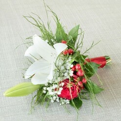 Seen from above upon a subtly textured, light beige linen reminiscent of stylish Hampstead interiors, this finely crafted bouquet captures the refined floral artistry of Golders Green. A brilliant white lily, fully opened and fragrant, forms the luminous focal point. Its smooth petals sweep outward with a gentle grace, each freckled in russet specks and sheltering cheerful yellow anthers-the essence of purity and quiet celebration. Nestled close, a lush red rose glows with velvety warmth, its petals a deep, romantic spiral. Bright, reflective red berries cluster within the greenery, their presence evoking the festive cheer and cozy winter walks found in local North End parks. Sprigs of delicate baby's breath lend a light, cloudlike softness, bridging the vibrant tones. To the left, a slender green lily bud leans contentedly, hinting at fresh growth and the promise of more blooms-echoing the garden charm of Golders Green's tree-lined avenues. Framing the composition, a lively blend of finely textured ferns and rich, broad leaves add lush depth and movement. The elegant stems are united with a satin red ribbon, tied with care. Altogether, this arrangement's harmonious palette and intricate textures evoke joyful events, heartfelt milestones, and the best of Golders Green floristry-crafted for meaningful occasions and same-day delivery.