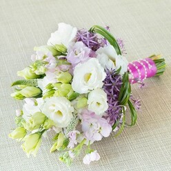 A radiant bouquet, hand-tied and thoughtfully designed, rests upon a subtly textured beige fabric-likely linen or burlap-bathed in gentle daylight that evokes the quiet calm of Golders Green mornings. Creamy white Lisianthus blossoms with their signature ruffled elegance take center stage, their satiny petals unfolding beside a scatter of unopened buds, while delicate pale lime accents suggest fresh promise. Generous clusters of lavender-pink stock add a soft, airy touch, reminiscent of blossoms found along the leafy borders of Golders Hill Park. Deep purple alliums introduce bold architectural notes, reminiscent of the creative vibrancy seen in the local galleries and cafes. Slender, looping greenery brings dynamic movement, echoing the arcs of nearby tree-lined avenues. At the bouquet's base, a vivid fuchsia satin ribbon provides a lively pop of color, secured with lustrous strands of white pearls-perfect for an elegant Golders Green wedding or celebratory breakfast. Every element, from soft pastels to dramatic purples and bright greens, suggests both serene romance and exuberance. This bouquet is more than a floral arrangement; it's a celebration of unity and artistry, capturing the timeless emotion of special moments shared in the heart of Golders Green.