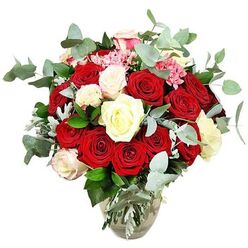 A richly detailed bouquet brimming with roses is shown in front of a spotless white backdrop, the kind of luxurious arrangement you might find gracing a Golders Green townhouse living room near Hampstead Garden Suburb. The flowers spill generously from a clear glass vase whose rounded base and narrower neck hold the stems in a neat vertical cluster, the water line visible and sparkling. At the centre, deep red and vibrant scarlet roses create a powerful core, their petals velvety and layered, some blooms just beginning to unfurl while others are open in wide, romantic spirals. Woven among them are white and creamy ivory roses, along with a few white blossoms tinged softly with blush pink at the tips, bringing light and a gentle, hopeful mood to the ensemble. Between the larger flowers, tiny sprays of bright pink filler blooms, perhaps miniature carnations or berry-like clusters, dot the arrangement with playful spots of colour and fine texture. Generous stems of eucalyptus, with their blue-green, coin-shaped leaves, arc gracefully around and between the roses, adding a silvery freshness and suggesting a subtle, calming scent. Deeper green leaves and touches of softer silvery foliage give depth to the base, ensuring the arrangement looks full, rounded, and impeccably composed from every angle. The overall impression is of abundant romance and contemporary style, a bouquet that feels equally suited to a Valentine's surprise arriving at a flat off Golders Green Road or a milestone celebration dinner near Brent Cross, brought to life by the practiced hands of a local florist who understands both classic beauty and modern London taste.