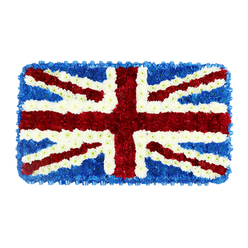 A meticulously crafted floral Union Jack, shaped into a perfect rectangle, rests against a pure white background, its colours and textures standing out with solemn clarity. The entire flag is composed of tiny, tightly packed blooms that form a dense, cushion-like surface, reminiscent of a ceremonial standard folded with care. Deep blue flowers create the outer field, their clustered heads forming a smooth yet softly rippled texture that frames the design, much like the calm, tree-lined streets around Golders Green. Over this blue base, the broad red St George's Cross is laid out with absolute precision, using vivid crimson blossoms whose velvety petals catch the light and appear gently raised. Interlaced with it, the thinner red diagonals of St Patrick's Cross cut confidently across the flag, each line edged with fine white borders. These white bands, made from crisp white chrysanthemums or similar flowers, also open up into the wider diagonal sections of St Andrew's Cross, which add a sense of light and space to the composition. Every junction where red, white, and blue meet is sharply defined, showing the steady hand and experienced eye of a professional florist used to preparing tributes for heartfelt services in local chapels and homes around Temple Fortune and Hampstead Garden Suburb. The overall impression is one of reverence and collective memory: a patriotic floral emblem intended for funerals, military commemorations, or national remembrance days. Though you cannot see their fragrance, the fresh blooms suggest a gentle, earthy scent, bringing a touch of natural comfort and quiet pride to a Golders Green farewell.