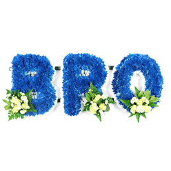 Framed against a stark white studio background, three freestanding letters form the word BRO, each letter built from a dense carpet of royal blue chrysanthemum blooms. The texture is plush and sculptural; every tiny floret contributes to a smooth, saturated surface while the white structural base peeks through in neat outlines around the inner curves and circular spaces. Each character is softened by a small cluster of pale ivory spray roses and tiny white fillers-possibly mini chrysanthemums or baby's breath-placed thoughtfully at the lower-left of the B and toward the lower-right of the R and O. Lush green elements such as broad salal leaves and lighter fern-like fronds punctuate the blue massing, introducing depth, movement, and an understated green scent. The overall lighting is calm and clinical, allowing the blue to read as serene and the creamy accents to glow, reinforcing an atmosphere of quiet remembrance. The composition is meticulous and dignified, a bespoke memorial piece often selected by families in Golders Green, Finchley and neighbouring Hampstead when they want a clear personal statement at a service. The combination of royal blue for solace and loyalty with cream roses for purity creates a visual language of consolation and connection, an intimate tribute to a brother or close friend that holds form, colour and sentiment in equal measure.