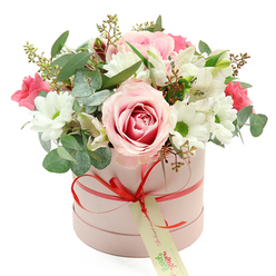 A refined and generously filled hatbox arrangement sits centrally against a bright white backdrop, its pastel tones and fresh greenery creating a sense of calm luxury. The container is a smooth, light pink cylindrical box, elegantly simple and reminiscent of high-end gifts carried from the shops around Golders Green station. Wrapped around its middle is a narrow, coral-red satin ribbon, shining softly and tied into a small, precise bow at the front. From the bow dangles a cream gift tag with green, handwritten-style lettering, suggesting a personal message and the care of a neighbourhood florist who knows local occasions well.   

The bouquet rises from the box in a rounded, dome-like shape, overflowing just enough to look abundant without feeling crowded. At the forefront sits a striking, fully opened rose shaded from pale pink at the edges into a deeper pink spiral at its heart, like a sunset over nearby Hampstead Heath. Surrounding this central rose, white daisy-like chrysanthemums with bright green centres bring a clean, cheerful brightness, their petals overlapping slightly to create soft, cushiony textures. Between them are slender stems of white alstroemeria, each blossom lightly speckled inside, adding a whisper of detail that rewards a closer look.   

Scattered throughout are smaller, rich fuchsia blooms-perhaps spray roses or lisianthus-that punctuate the softer colours with lively energy. Lush greenery frames everything: rounded, silver-green eucalyptus leaves curve gently, darker foliage anchors the arrangement, and tiny green buds contribute a sense of freshness and movement. The composition feels ideal for marking tender moments-Mother's Day, an anniversary in NW11, or a quiet 'thinking of you' sent to a friend living off Finchley Road-capturing both elegance and heartfelt emotion in one thoughtfully curated hatbox.