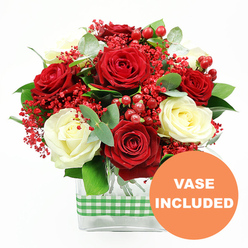 This image presents a lush, carefully composed bouquet that seems to glow with warmth against a clean white backdrop, much like a welcoming window display on a winter evening in Golders Green. The arrangement is dominated by full, velvety red roses, their petals layered in deep crimson whorls, partnered with smooth, creamy ivory roses that soften the palette and introduce a note of quiet elegance. Nestled between these larger blooms are delicate sprays of tiny red filler flowers, similar to dyed gypsophila, which create a fine, frothy veil over the composition, suggesting a dusting of festive confetti. Glossy, rounded red hypericum berries punctuate the design, adding a jewelled sheen and tangible texture that would catch the eye of anyone strolling past a florist on Finchley Road. Around and beneath the flowers, eucalyptus and other fresh green foliage curve and unfurl, their leaves ranging from deep green to muted blue-green, adding depth, movement, and a subtle sense of fragrance. The bouquet is arranged in a sleek, rectangular clear glass vase, allowing the orderly stems to be seen and giving the arrangement a tidy, contemporary feel that would complement modern apartments and traditional homes across Golders Green alike. A green and white gingham-style ribbon wraps horizontally around the middle of the vase, its charming country pattern hinting at rustic kitchens and relaxed family gatherings. In the bottom right corner of the photograph, a bright orange circular graphic with crisp white lettering reads "VASE INCLUDED," clearly signalling that the flowers arrive ready to display as soon as they are delivered. Overall, the scene conveys a mood of celebration and affection, suitable for anniversaries, Valentine's surprises, or simply brightening a home near Golders Hill Park with fresh, locally arranged blooms.