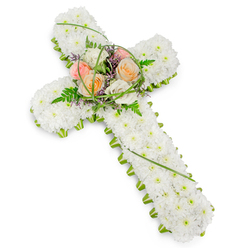 A beautifully composed sympathy cross is shown against a bright, uncluttered white backdrop, allowing every floral detail to stand out with clarity and grace. The cross itself is fully padded with countless small white chrysanthemums, each flower carefully placed so that no gaps interrupt the soft, cushion-like surface. Their bright, clean petals form a calm, snowy expanse that suggests innocence and eternal rest. Running along the outer edge of the cross is a fine border of fresh green foliage and structure, hugging the arms and base and helping the silhouette remain sharp and well-defined. At the centre, where the beams of the cross intersect, a raised spray of larger blooms becomes the emotional focal point of the design. Here, several roses open in gentle, comforting shades of pastel peach, warm blush rose, and creamy off-white, their layered petals catching the light and drawing the eye inward. Wispy blades of bear grass curl and loop over the roses, while feathery fern fronds extend outward, softening the edges of the spray. Tiny purple filler flowers peek through the greenery, introducing a subtle hint of colour that enriches the overall palette without overpowering the tranquillity of the white base. The arrangement feels quiet yet expressive, much like a thoughtful farewell offered in one of Golders Green's chapels or synagogues after a local service. This floral cross would sit beautifully beside a coffin, at the front of a memorial gathering, or near a resting place, providing a symbol of faith, remembrance, and gently held memories for families in and around Golders Green.