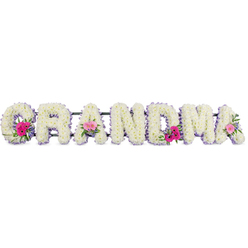 This image shows a beautifully crafted funeral flower tribute that spells out the word "GRANDMA" in large, upright letters, each one luxuriously covered in pristine white chrysanthemums. The blossoms are densely arranged so that no gaps in the letters are visible, forming a soft, cushion-like surface with countless little petals overlapping like feathers. Around the edge of every letter runs a slim lilac ribbon, the pastel purple trim giving each shape a clear outline and adding a calm, graceful contrast to the snowy white blooms. Three focal clusters of colour are woven into the design: on the first "G," one of the central "A"s, and the last "A," compact posies of bright pink gerbera daisies and soft pink roses sit proudly on top of the chrysanthemums. These accent flowers create joyful bursts of pink against the white canvas, while sprigs of fresh foliage and small leaves lend touches of green that feel soothing and natural, like a walk through Golders Hill Park on a quiet afternoon. The tribute lies on a flat surface, likely prepared by experienced florists at Flowers Golders Green before a service or family farewell. The overall mood of the piece is tender and reverent-an elegant floral message that feels both strong and gentle, perfectly suited to honouring a grandmother whose presence once warmed family gatherings in Golders Green, Temple Fortune, or nearby streets. The composition balances purity, softness, and subtle colour, capturing the essence of remembrance and enduring love.