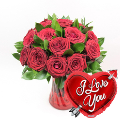An opulent cluster of deep red roses rises from a ruby-tinted glass vase, creating a bold and passionate statement against a spotless white backdrop. The bouquet holds approximately one dozen or slightly more roses, each bloom rich with layered, velvety petals that soften towards the edges, showing subtle curves and ruffles as they open fully. Some flowers are in generous, open bloom, while others are still gently unfurling, suggesting love that is both established and still unfolding. Among the roses, glossy dark green foliage provides contrast, its smooth, slightly waxy leaves creating pockets of shadow and depth that make the crimson tones glow even more intensely. The vase itself is made of red glass with delicate fluted or ridged detailing, its translucent surface catching the light and sending a warm, ruby shimmer into the water inside. In the lower right corner of the image, overlapping the base of the bouquet, a bright, metallic red heart-shaped foil balloon leans forward, as if drawn toward the roses. The balloon's surface is mirror-smooth, with highlights reflecting along its rounded edges, and across its centre, in flowing white script, the words I Love You appear clearly, accompanied by a silver Cupid's arrow that seems to pierce the heart from corner to corner. This romantic arrangement feels like something a Golders Green florist would prepare for a proposal in a cosy restaurant off Golders Green Road or a surprise anniversary gift delivered to a home near Hampstead Garden Suburb. The simple white background ensures every detail-the red, the green, the glossy balloon-stands out, evoking warmth, affection, and a sense of celebratory romance.