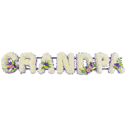 A horizontal floral name tribute spelling GRANDPA is captured in crisp detail against a plain white backdrop, as if set on a workbench in a careful florist's studio near Golders Green Road. Seven bold capital letters sit in a straight line, supported by a slim, dark green metal bar that discreetly anchors the arrangement while allowing the flowers to remain the focus. Each letter is built from a thick cushion of white chrysanthemums or similar multi-petalled blooms, packed so densely that no gaps show, giving a velvety, almost quilted texture. Running around the rim of every letter is a neat edging of small lilac and soft purple flowers, providing a delicate frame that picks out the curves and angles. Within selected letters, the florist has woven in richer accents: the 'G' and first 'A' hold sprays of lily-like white flowers, surrounded by pink and fuchsia blossoms and deeper purple tones, while touches of glossy green foliage break the white and add natural movement. The 'D' and another 'A' echo these clusters, including tiny green button chrysanthemums that offer a bright, hopeful spark amidst the calm palette. The soft, even lighting emphasises the freshness of each petal, the cool whites and gentle purples suggesting quiet reflection rather than drama. The overall feeling is one of tenderness and respect, a dignified way to say goodbye to a much-loved grandfather whose life might have included strolls through Golders Hill Park or visits to the local cafés by the station. Crafted with the care you would expect from experienced Golders Green florists, this funeral flower arrangement becomes a wordless message of gratitude, remembrance, and enduring family love.
