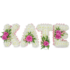 This floral name tribute presents KATE in sculpted, freestanding letters formed from a dense carpet of small, immaculate white chrysanthemums that glow softly against a pristine white backdrop. A whisper of pale pink satin ribbon outlines each letter's inner edge, lending a refined yet gentle contour that plays with the light and frames the composition like a framed portrait. Strategic clusters of hot pink roses-deep, lively fuchsia-act as visual anchors: two open roses decorate the left side of the K, a single bold rose sits within the lower arc of the A, the base of the T is punctuated by a single vivid bloom, and the E carries a tandem of roses on its right-hand face. Lush green accents-fern fronds, ruscus and variegated foliage-slide quietly beneath and between blooms, while fine pink buds and delicate alstroemeria-like sprays provide soft, garden-fresh detailing. The chrysanthemums' plush texture creates a cloud-like surface, their compact packing forming a continuous white field that contrasts with the roses' velvety petals. Faint shadows beneath the letters emphasize the arrangement's three-dimensional presence, suggesting it is freestanding and ready for careful placement at a memorial table or reception. A florist familiar with Golders Green and the nearby Finchley Road will recognise this style as an elegant, personal tribute often requested for funerals, memorial services, or intimate celebrations of a life-reserved in tone, tactile in texture, and quietly expressive.