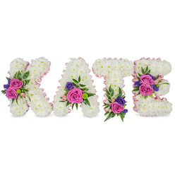 A refined memorial display reads "KATE" in four freestanding letters, each letter formed from a dense, uniform carpet of pure white chrysanthemums whose tiny heads create a pillowy, tactile surface. A narrow satin ribbon in a pale, blush-pink outlines every outer and inner edge, giving the letters a soft, luminous definition like a whispered greeting. The white theatre of blooms is interrupted by artful clusters of colour: deep fuchsia-pink roses provide a bold, velvety centre, while smaller pastel pink spray roses cascade more delicately around them. Sprigs of deep purple or navy-toned filler - likely statice or compact lisianthus - give each cluster an anchoring contrast, and fresh greenery such as fern fronds and eucalyptus leaves adds airy movement and a cool, textured backdrop. Placement is intentional and balanced: the diagonal of the "K" bears a prominent arrangement, the lower right of the "A" nestles a rounded spray, the base of the "T" carries a comforting cluster, and the "E" is adorned twice - both at its middle bar and lower arm - creating a visual rhythm across the name. The composition is photographed on a clean white field that emphasizes colour and craft; the overall mood is one of dignified affection, a floral tribute often chosen for funerals, memorials, or graveside remembrances in Golders Green and nearby Hampstead Garden Suburb. The design reads as both personal and serene, a tactile lettered homage that speaks when words feel inadequate.