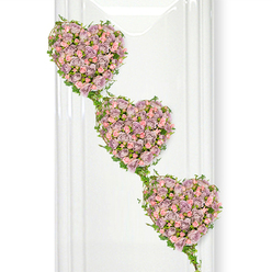 A refined floral tribute of three interlinked hearts takes centre stage in this image, each one carefully shaped from a dense tapestry of roses that speak softly of devotion and remembrance. The hearts descend in a gentle diagonal from the top left corner, creating a flowing, almost musical rhythm, as if each heart were a verse in the same loving story. Every heart is filled edge to edge with muted lavender and dusky mauve roses, interspersed with paler pastel pink blooms that catch the light and lend a tender, comforting warmth. The petals look velvety and layered, some blooms fully open and generous, others still cupped, suggesting both memories cherished and moments that will always remain close. Slim strands of ivy and fresh green foliage trace the outlines of the hearts, curling around their contours and then reaching out to link one to the next, like hands gently joined. The leaves, a clear, healthy green, introduce a sense of nature and renewal, reminiscent of a walk through nearby Golders Hill Park when the borders are full and flourishing. The arrangement is shown against a clean white backdrop with faint vertical reflections on the left, hinting at glass or polished stone and evoking the stillness of a chapel or a peaceful funeral parlour in Golders Green. The overall mood is tranquil and sincere, ideal for honouring a loved one's life, offering comfort at a service, or sending a message of unbroken love and gratitude from family and friends.