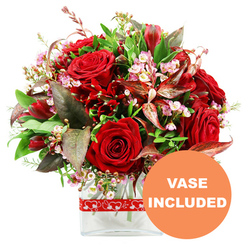 A luxurious dome-shaped bouquet filled with rich red blooms is photographed against a pure white background, as if displayed in the window of a Golders Green florist on a bright morning. Dominating the composition are velvety red roses, some in tight buds promising days of beauty, others fully unfurled to reveal their layered petals and evocative depth of colour. Threaded between them are clusters of deep crimson flowers resembling alstroemeria, their multiple petals curling and folding to create an intricate counterpoint to the classic roses. Interspersed throughout the arrangement, tiny sprays of pale pink and white filler blooms, akin to waxflowers, weave a light, cloud-like texture that gently lifts the heavier tones. Encircling this heart of crimson is a lush array of greenery: glossy dark leaves, softer green fronds, and distinctive burgundy and reddish-brown foliage, some with elegant veining that catches the light. These darker leaves lend a sophisticated, almost dramatic frame, much like the quiet, tree-lined streets just off Finchley Road framing the bustle of Golders Green. The bouquet is carefully placed in a clear rectangular glass vase with softly rounded edges, its transparency allowing a glimpse of the green stems and water, adding a sense of freshness and modern simplicity suited to contemporary flats and townhouses. A vivid red ribbon decorated with small white heart outlines is wrapped around the vase, subtly suggesting romance and affection without feeling overly ornate. In the lower right corner, a circular orange badge stands out, carrying the bold white text VASE INCLUDED, instantly signalling that this arrangement arrives ready to display. The overall mood is one of passionate elegance, ideal for celebrating an anniversary, surprising a partner after a stroll through Golders Hill Park, or sending a heartfelt romantic gesture across Golders Green.