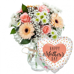 Set against a crisp white background, this Mother's Day scene feels like a quiet moment in a Golders Green flower studio, where every stem has been placed with care. A clear glass vase holds a generously rounded bouquet, its neck cinched with a simple loop of natural twine that gives the arrangement a relaxed, homespun charm. The flowers themselves glow in soft pastel tones: peach-pink roses open wide to reveal elegantly layered spirals of petals, their gentle blush colour evoking calm weekend walks through the gardens of Golders Hill Park. Nestled beside them, bold peach gerbera daisies, each with a deep chocolate-brown centre, introduce a joyful, sunlit energy. Between these focal blooms, airy sprays of white gypsophila drift like tiny clouds, while petite white chrysanthemums with subtle apple-green centres build a cushion of texture and volume, recalling small cottage flowers gathered from local gardens off Finchley Road. Fresh green foliage threads throughout the design, adding contrast and framing the lighter blooms with a leafy, natural border. Just behind the flowers, almost hidden, sits a small white card with hints of pink and green writing, suggesting a personal Mother's Day dedication. In front and slightly to the right of the vase, a glossy heart-shaped foil balloon becomes a playful companion to the bouquet. The balloon's white surface is dotted with glimmering gold and silver confetti spots, and at its centre lies a peach heart carrying the message HAPPY Mother's DAY in bold black type and elegant script. Together, the flowers and balloon form a warm, celebratory display, perfect for a Mother's Day delivery across Golders Green and the surrounding neighbourhood.
