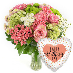 This image shows a luxurious Mother's Day flower and balloon gift set, styled as a clean product photograph on a bright white background, much like the displays you might see in a Golders Green flower shop before morning deliveries go out. A clear glass vase holds a lush, domed bouquet overflowing with fresh blooms in pink, coral, lime green and white. The eye is drawn first to several generous pink roses, whose petals deepen in colour toward the centre and soften to paler edges, suggesting tenderness and affection. Threaded around them are clusters of tiny coral-pink blossoms, similar to kalanchoe, forming dense, starry heads that bring texture and a slightly modern twist. Lime-green button chrysanthemums and velvety chartreuse florets create a playful contrast, their rounded forms and frilly surfaces echoing the lively character of nearby Golders Hill Park in spring. Between these sit delicate white daisy-like spray chrysanthemums and a few smaller, spiky coral blooms, giving the arrangement a light, airy movement. Dark green foliage cups the flowers and spills gently outward, grounding the brighter tones and framing the composition. The stems, neatly cut and immersed in clear water, are visible through the transparent vase, reinforcing the sense of just-picked freshness, perfect for same-day delivery around NW11. To the lower right, a shiny heart-shaped foil balloon completes the gift. Its white surface is dotted with irregular spots of gold, silver and soft peach, and at the centre a bold peach-coral heart bears the message HAPPY Mother's DAY in stylish black lettering. The overall mood is celebratory yet heartfelt, capturing the feeling of bringing a smile to a mum's face in Golders Green, Temple Fortune or along Finchley Road on Mother's Day.