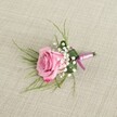 A single blush pink rose boutonniere rests on a softly textured beige linen cloth, photographed from a slightly elevated angle so you can almost feel yourself leaning over a dressing table in a Golders Green townhouse before a wedding. The rose is semi-open, its velvety petals spiralling gently outward from a tighter heart of pale, powdery pink into edges that deepen into a richer, romantic rose-pink. Each petal catches the soft, diffused light, which seems like an overcast North London afternoon, revealing delicate shading and a faint sheen that suggests just-misted freshness. Around the base of the bloom, clusters of creamy white baby's breath form little clouds of tiny star-like flowers, their rounded buds and open florets adding a light, airy halo that softens the composition. Fine, grass-like foliage in bright, emerald green fans out from behind the central rose, its slender blades slightly curved, adding movement and a hint of wild garden charm you might see walking through nearby Golders Hill Park. A broader, deeper green leaf is neatly folded and wrapped around the stems, giving the boutonniere a tailored, structured base reminiscent of a well-cut suit lapel. The stems are carefully bound with a satin ribbon in two harmonious tones: a soft lavender-pink that blends with the rose, and a deeper magenta-purple running alongside, tied in a modest, secure knot without trailing ends. A small, silver-toned pin sits subtly at the base, indicating readiness to be fixed to a groom's or prom guest's lapel. The natural linen background, with its visible weave and warm neutral tone, provides a calm, understated backdrop that lets every petal, sprig, and ribbon fold stand out, capturing the quiet anticipation of a formal celebration in Golders Green.
