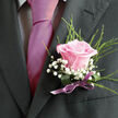 The image presents a close, intimate view of formal attire, the camera lingering over the textures and colours that mark a special occasion in Golders Green. A dark charcoal or black suit jacket fills most of the frame, its left lapel crisply tailored and catching just enough light to reveal its smooth weave. Beneath it, a pale dress shirt with a fine gingham check peeks through, providing a gentle pattern that supports rather than competes with the strong vertical of a vivid magenta-pink tie. The tie, perfectly knotted and silky in finish, draws the eye downward to the true star of the composition: a meticulously arranged boutonniere. At its centre, an exquisite light pink rose opens in graceful spirals, petals deepening to a warmer rose tone near the heart and fading to a soft blush along the edges. Surrounding this bloom, tiny white baby's breath flowers create a delicate haze, their miniature petals catching the light like a sprinkling of frost. From behind, wisps of fine, bright green foliage arc outward, lending height and an organic, slightly whimsical touch, reminiscent of fresh greenery found in local gardens off Finchley Road. The stems are concealed within a broad, glossy green leaf, cinched with a narrow, satiny purple ribbon tied into a tidy bow that rests just above the jacket's breast pocket. The colours of rose, ribbon, and tie harmonise beautifully, suggesting a carefully coordinated outfit for a wedding, engagement party, or gala nearby, with the boutonniere embodying affectionate celebration and the promise of a memorable day.