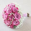 A radiant, hand-tied bouquet, perfect for Golders Green weddings or romantic occasions, rests invitingly on a soft, light neutral fabric, reminiscent of the tranquil interiors of local venues. At the heart of the bouquet are approximately a dozen lush roses displaying a mesmerizing spectrum of colour-from dusty, blush-pink centres to vivid magenta rimmed petals that catch the eye even in gentle daylight. Wispy fronds of baby's breath (Gypsophila) form an ethereal frame around the roses, their snow-white, cloud-like clusters offering a striking contrast and amplifying the overall volume and softness of the bouquet. Each bloom is impeccably fresh, clustered in a classic, rounded arrangement that hints at the skilled touch and artistry of Golders Green's esteemed florists. The tightly bound stems are gracefully wrapped in a glossy, pale pink satin ribbon, completed by a delicate spiral strand of small, shimmering pearls-both practical and elegant, providing a comfortable grip for heartfelt celebrations. The fresh, green-brown stems extend just beyond the ribbon's end, signaling an authentic, hand-crafted creation. Soft, ambient light plays across the petals and gypsophila, highlighting every nuanced detail and lending the bouquet a serene, almost dreamlike quality. Thoughtfully composed, this stylish arrangement is a perfect choice for local brides, anniversaries, or anyone seeking a sophisticated romantic gesture steeped in Golders Green's tradition of floral excellence and care.