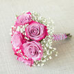 Resting on a neutral, woven beige background that recalls the calm interiors of many Golders Green homes, this compact bouquet showcases a careful balance of drama and softness. The eye is drawn first to the roses, each head a beautifully formed spiral of petals. Their centres glow with a muted lavender or soft lilac tone, while the outer edges burst into an intense band of dark fuchsia, almost magenta pink, giving the blooms a painted, two-toned effect. These roses are gathered closely so that their rounded shapes form a single, opulent cluster. Threaded generously between them is white baby's breath, its countless pinhead blossoms forming a delicate haze that frames the roses and introduces a sense of air and innocence. The contrast between the crisp white gypsophila and the rosy purples is gentle yet eye-catching, like the changing light over nearby Hampstead Heath on a clear evening. The stems are bound tightly in a sleek, deep purple satin ribbon, the fabric folded with precision. Winding around this handle is a string of luminous faux pearls, coiling in a careful spiral that adds texture and a nostalgic bridal charm, ideal for weddings at local venues or registry offices near Golders Green Underground. The bouquet is angled diagonally across the image, with the pearl-wrapped stems pointing to the right, suggesting movement and readiness, as though it has just been set down between photographs. It feels intimate and personal, a handcrafted floral accessory suited to bridesmaids, prom celebrations, or romantic gestures, freshly arranged for local delivery by an experienced NW11 florist.