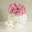 On a clean, light surface, a contemporary wedding arrangement radiates gentle happiness, much like a calm morning in Golders Green before the day's celebrations begin. The eye is drawn first to a compact dome of lush pink roses, each bloom perfectly open yet still full of life, petals curling outwards in delicate spirals. Their colour moves softly from pale, milky pink at the base of each petal to a deeper, almost raspberry hue along the edges, giving the flowers a hand-tinted look. Nestled between the roses, sprigs of white gypsophila-baby's breath-rise like tiny clouds, softening the lines and lending a whispery texture. The bouquet is cradled in an all-white, hand-woven spherical container, formed from curved strips that loop and twist around each other in a lattice. This orb feels both sculptural and airy, its open spaces allowing glimpses of shadow and light, as though the flowers float above the table. The material appears smooth and slightly matte, echoing the clean, fresh aesthetic often seen in chic North London wedding venues. At the base, two small, similarly woven decorative spheres sit to one side, adding balance and a minimalist, design-led touch. There is no clutter around the piece; just this single, confident arrangement that suggests calm elegance and refined joy. It is easy to imagine it gracing a wedding breakfast table near Hampstead Garden Suburb or welcoming guests at a Golders Green registry ceremony, offering a gentle fragrance of roses and the quiet promise of a beautiful day.