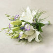 This fresh, ethereal bouquet is presented on a pale beige linen surface and captures the hush of an early summer morning in Golders Green. White lilies take centre stage, their trumpet-like flowers opening to luminous, creamy throats and prominent golden stamens dotted with tiny specks, while nearby green buds sit ready to bloom. Interspersed are lisianthus blooms whose petals carry a delicate lavender-purple blush along their ruffled edges, offering a restrained yet playful contrast to the pure whites. Sprays of airy, fern-like greenery mingle with broader glossy leaves to create layered depth and a gentle sense of movement, as if a soft breeze from the nearby Finchley Road had just passed by. Scattered tiny clusters of baby's breath add an almost cloud-like lightness, lifting the composition away from heaviness. The stems are gathered and finished with a crisp white satin ribbon toward the right, revealing a thoughtful, hand-tied construction that lies low and easy for holding or placing on a tabletop. Soft directional light sculpts subtle shadows and brings out the linen's texture, enhancing the bouquet's tactile qualities and hinting at the fresh floral scent. Ideal for intimate gatherings, a refined gift, or a wedding bouquet for someone who prefers quiet elegance, this arrangement feels both restorative and sophisticated - a floral moment that reflects the calm pockets of greenery tucked into the Golders Green neighbourhood.