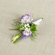 A finely crafted wedding boutonniere lies against a softly textured light beige linen background, its calm neutrality allowing every petal and leaf to stand out with clarity. Two white lisianthus blooms form the heart of the design, their petals gently ruffled and edged in a deep, velvety purple that pools toward the tips like watercolour ink. Around them, tiny white filler flowers, similar to baby's breath, float like small clouds, giving an airy softness and a sense of breath between the bolder colours. A slender vertical spike of tightly clustered, bell-shaped muscari flowers introduces a rich grape-purple note, their bead-like texture adding quiet intrigue. Fresh greenery, including fine, feathery fern-like fronds and a few broader leaves, cradles the blooms, creating a light, meadowy feel that could just as easily belong to a walk through nearby Golders Hill Park on a cool spring morning. Two pale green buds sit just off-centre, still closed, evoking that moment before a ceremony begins, when anticipation hangs gently in the air. At the base, the stems are neatly gathered and wrapped in crisp white satin ribbon, the fabric smooth and slightly lustrous, tied so precisely it feels ready to be pinned to a groom's lapel before a Golders Green synagogue wedding or a smart reception near the station. The overall mood is refined but not fussy, a small cluster of flowers that speaks in quiet, confident tones, ideal for anyone wanting a subtle, elegant accent to their formal suit for a wedding, prom, or special evening celebration in North West London.