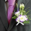 A close-up photograph captures the upper torso of a man dressed for a smart city wedding in Golders Green, the camera lingering lovingly over every refined detail. He wears a dark charcoal grey suit jacket, perfectly tailored so the lapels sit clean and sharp, framing a pale grey checkered dress shirt beneath. At the centre of the ensemble, a vibrant magenta silk tie glows with a soft sheen, its knot pulled neatly into place, adding a confident burst of colour that feels quietly celebratory. Pinned to the left lapel is a meticulously crafted boutonniere, the true focal point of the image. The main bloom is a white lisianthus flower edged in deep purple, its ruffled petals opening like a delicate watercolor, with a bright yellow centre that draws the eye. A second lisianthus bloom nestles alongside the first, supported by several slender, unopened white buds that suggest new beginnings. Rising gracefully behind them is a single spire of indigo-purple veronica, its tiny florets forming a vertical line that adds height and distinction. Around these feature flowers, fine sprays of baby's breath and airy greenery create an almost meadow-like softness, with feathery textures and looping strands of decorative grass adding a contemporary twist. A crisp white satin ribbon is tied into a small, precise bow at the base, securing the stems and lending a touch of traditional elegance. The lighting is gentle but clear, catching the satin of the tie and the satin sheen of the foliage, making the whole buttonhole feel like a fresh, fragrant moment of joy, perfect for a groom heading to a ceremony near Golders Hill Park or one of Golders Green's local synagogues.