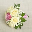 A charming, compact bridesmaid's bouquet rests softly on a subtly textured, cream-colored linen-its arrangement a picturesque nod to Golders Green's tradition of refined weddings. Enveloping the center are five creamy roses, their petals unfolding in gentle, spiraling layers and filling the composition with a sense of warmth and serenity. Nestled slightly off to one side shines a single deep pink rose, its magenta hue a striking twist of colour, like the unexpected charm of a local garden hidden just off Golders Green Road. Whisper-light clusters of pure white gypsophila (baby's breath) thread through the bouquet, lending a delicate lacework of texture and brightness. Sparse, glossy green leaves peek from underneath, their fresh sheen underscoring the seasonal vibrancy of the arrangement. The bouquet's stems, gathered and neatly bound by a vivid pink ribbon, echo the magenta rose above while providing a welcoming tactile finish. Wrapping the handle is a spiraling trail of luminous white pearls, their subtle glimmer enhancing the bouquet's romantic aura. Illuminated by gentle, natural Tonal light, the layered petals and greenery create a portrait of local floral artistry, perfect for intimate nuptials, celebratory gatherings, or elegant event delivery across Golders Green. The bouquet evokes the gentle excitement of special occasions-a timeless piece imbued with local character, ready to bring a touch of romance and serenity to any Golders Green celebration.