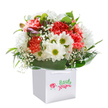There is an undeniable delight woven into this bouquet, springing alive with a vibrancy that reflects the cheerful spirit of Golders Green's leafy streets. Coral and red carnations dominate, their petals lushly layered-so soft and abundant, you can almost feel their velvety touch just by looking. Crisp, white daisies bloom beside them, bright centres hinting at the optimism of a sunny morning near Golders Hill Park. The arrangement practically glows as tiny baby's breath flutters amongst the bolder blooms, infusing the whole bouquet with gentle innocence and airy lightness. Deep green leaves encircle everything, glossy and structured, holding every stem in perfect balance. Subtle purple-tinged buds nestle among the open flowers, adding a playful hint of mystery. All of these are thoughtfully arranged in a spotless square white gift bag, the white ribbon handles arching above, ready for easy presentation to a friend, a neighbour on Hoop Lane, or as a surprise for a family celebration. Each stem reveals the care and expertise of a local florist, transforming flowers into a heartfelt message of joy and renewal. The image radiates with the freshness of a just-completed bouquet, destined to uplift-a perfect gift for birthdays, milestones, or simply to bring a smile to someone special in Golders Green.