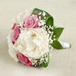 Laid horizontally against a pale, slightly textured fabric that suggests finely woven linen or cotton, this bridal bouquet seems to float in a pool of soft light, much like a quiet early morning in Golders Green before the shops open along Finchley Road. A large creamy white peony dominates the centre, its petals layered in abundant ruffles that form a plush, rounded bloom, inviting the imagination to feel their silky softness. Encircling it, several vivid fuchsia-pink roses add structure and contrast, each bud tightly packed and elegantly spiralled, conveying classic romance and poised anticipation. Throughout the bouquet, dainty clouds of white gypsophila are threaded with care, their tiny blossoms forming a lace-like veil that softens the strong rose tones and complements the peony's gentle cream. Deep, polished green leaves curve and fold around the flowers, their broad surfaces catching the light and offering a cool, botanical frame that makes every petal stand out. The stems are gathered with precision and bound in smooth white satin ribbon, its subtle sheen echoing the refined feel of a wedding celebration at a Golders Green venue or a reception near Hampstead Garden Suburb. The lighting is mellow and diffused, highlighting edges and textures while keeping shadows gentle, so the whole arrangement glows with understated elegance. It looks like a bouquet freshly prepared by an expert florist, ready for delivery to a bride who wants a timeless, romantic arrangement that feels natural, serene, and beautifully suited to a North London love story.