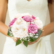 This image shows a refined bridal bouquet resting as though just placed down after a ceremony in Golders Green, its carefully chosen flowers arranged in a classic, rounded shape. At the centre, creamy-white peonies unfold in plush layers, their petals silky and full, reminiscent of the softest fabric on a wedding gown. Surrounding them, roses in tones of dusty pink and cool lavender add a sophisticated colour contrast, each bloom tightly constructed yet on the verge of opening further, creating a sense of anticipation and gentle movement. Tiny clouds of gypsophila weave between the larger flowers, their minute white blossoms catching the light like a scattering of confetti outside the registry office on Finchley Road. The deep green foliage creates a smooth, polished collar around the bouquet, the leaves slightly waxy and arching just enough to frame the blooms without overpowering them. The stems are gathered in a traditional hand-tied fashion, implying the secure, comfortable grip of a bride making her way from a ceremony in nearby Temple Fortune to a reception with close family and friends. Soft daylight falls across the bouquet, revealing subtle shifts in tone from ivory to blush to lilac, and highlighting the textures-from the velvety rose petals to the feathery baby's breath. The composition feels intimate and contemplative, like a quiet moment away from the bustle of Golders Green Road, capturing the calm happiness and graceful romance of a wedding day bouquet arranged with care and long-practiced skill.