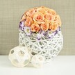 A tastefully composed wedding-style table arrangement rests on a pristine white surface, its reflection faintly shimmering beneath the blooms in a way that recalls a carefully laid dining table in a Golders Green home prepared for guests. The centerpiece is dominated by a rounded dome of peach and apricot roses, tightly massed so that their velvety petals form a near-perfect sphere. Hints of blush, salmon, and soft orange intermingle across the rose heads, creating a warm, inviting colour story that suggests romance without excess. Supporting this upper canopy of roses is a graceful collar of white lisianthus, whose petals are finely edged in vibrant violet or purple, adding a refined accent of colour that draws the eye around the base of the dome. Among them, delicate sprays of white baby's breath provide airy texture, filling in small gaps and softening the transitions between blossoms. These flowers are gathered in a bulbous white container, intricately woven from rattan or similar natural fibers, its open, basket-like lattice giving the design a light, handcrafted feel. The bright white of the vessel enhances the freshness of the flowers, while its rounded shape echoes the curve of the rose dome above. In front, two decorative woven spheres, crafted from pale beige wooden strips, rest casually on the same white surface, one larger, one smaller, adding a touch of sculptural interest that mirrors the container's texture. A soft beige fabric backdrop, subtly textured and gently lit, frames the entire composition with warmth. The overall impression is of calm elegance and thoughtful design, ideal for wedding tables, engagement parties, or intimate celebrations throughout Golders Green and nearby neighbourhoods.