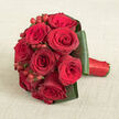 A compact, perfectly rounded bouquet of red roses rests on a softly textured beige fabric, its warm, neutral tone similar to natural linen laid across a dining table in a quiet Golders Green townhouse. The roses themselves are full and velvety, petals layered in tight spirals that deepen from bright scarlet at the outer edge to sultry burgundy in the heart. Some blooms are slightly more open than others, giving the arrangement a sense of movement and life, as though it has just been lifted from a bride's hands at the end of a ceremony near Golders Hill Park. Tucked among the roses are clusters of small, glossy hypericum berries in matching red, their smooth, rounded shapes catching the light and adding a playful, tactile accent to the lush, soft petals. Enveloping the base, broad dark green leaves have been folded with precision, forming a structured collar that holds the bouquet in a defined, contemporary silhouette while still feeling romantic and generous. The handle, visible at one side, is wrapped in rich red satin ribbon, its sheen echoing the berries and tying the whole design together with an elegant finish. Photographed from a slightly elevated viewpoint, every detail of the craftsmanship is clearly visible: the careful spacing of each rose, the rhythm of berry clusters, the crisp lines of green foliage. The calm beige background ensures that the deep reds and greens appear even more intense, making this arrangement feel like a heartfelt declaration of love, ideal for weddings, anniversaries, or a meaningful same-day delivery across Golders Green.