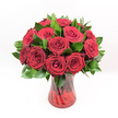 A studio-style photograph shows a classic, romantic bouquet of deep red roses standing alone against a pure white background, every detail crisp and clear. Around a dozen or slightly more velvety crimson blooms cluster together in a rounded dome, each flower perfectly formed, with spiralling petals and tightly layered centres that look ready to unfurl further. The roses are rich and saturated in colour, closer to burgundy at their depths and glowing brighter red where the light touches their outer petals, giving the arrangement a sense of movement and life. Between the blooms, glossy dark green foliage, likely Ruscus, peeks through in small, pointed leaves that frame the flowers and add fullness and contrast. The bouquet is gathered neatly into a translucent ruby-red glass vase with a subtly ribbed or swirling texture. The vase has a gentle hourglass shape with a flared rim and stable base, and the light catches its surface, creating soft reflections and highlights that echo the richness of the petals. Through the tinted glass you can see the darker lines of the rose stems, faintly crossing and tangling in the water, which grounds the airy blooms in something organic and real. The lighting feels like bright midday clarity, the sort you might find drifting in through a Golders Green shop window, revealing every curve and fold without harsh shadows. Altogether, the arrangement radiates passion, intimacy, and ceremony-ideal for a Valentine's Day surprise, an anniversary in one of the quiet streets off Finchley Road, or any grand gesture of love made more special by an expert local florist.
