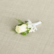 Seen in gentle diagonal composition on a light, subtly textured beige weave, this boutonniere centres on a pristine ivory rosebud whose layers of soft, creamy petals unfurl with delicate symmetry. Encircling it, tiny clusters of pure white gypsophila create a fine, cloud-like veil that lifts the eye and softens the silhouette, while slender, feathery sprigs of dark green foliage - reminiscent of evergreen or cedar - provide a cool, textural contrast. Deeper, broader green leaves sit behind the bloom, offering structural support and a velvety backdrop that emphasizes the rose's luminous cream. The stems are precisely bound with a narrow white satin ribbon, finished in a small, simple bow that reads as both ceremonial and quietly refined; the satin's gentle sheen catches the studio light without drawing attention away from the flowers. Resting on the woven fabric, the boutonniere conveys a faint, fresh fragrance: a whisper of petals and green sap that suggests an early spring morning. This wearable floral accent, typically pinned to a lapel, carries traditional symbolism of purity and new beginnings and is an elegant choice for grooms, ushers, or guests at weddings and formal occasions. Described with the calm confidence of a florist who knows the narrow lanes around Finchley Road and the leafy calm of Hampstead Garden Suburb, the image balances craftsmanship and sentiment, making it useful for wedding planning guides, floral styling advice, or local delivery information in Golders Green.