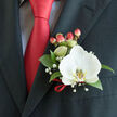 This close-up captures a carefully composed boutonnière pinned to the left lapel of a dark charcoal suit, where a single white Phalaenopsis orchid becomes the quiet hero of the arrangement. The orchid's wide, creamy petals sit softly against the suit's structured fabric, the orchid's yellow-and-white patterned lip offering a focused point of warmth against a sharply knotted red satin tie and a light-blue, grid-patterned dress shirt just visible beneath the jacket. Deep green foliage provides a steady, living backdrop; each leaf is glossy and firm, lending weight and form to the small cluster of baby's breath that drifts like a soft mist around the central bloom. Rounded hypericum berries, in a coral-to-red blush, echo the tie's vibrant tone and bring a tactile roundness that contrasts with the orchid's sleek curves. A fine red satin loop secures the stems, its sheen mirroring the tie and adding a considered finish that speaks of a florist's practiced hand. The lighting is gentle and natural, suggesting early afternoon or soft indoor illumination typical of a Golders Green reception or a photo taken near Golders Green Road. The overall impression is of careful restraint and joyful refinement - an emblem of ceremony and affection suited to weddings, anniversaries, or formal gatherings in the area, crafted to maintain freshness and subtle scent throughout the day.