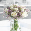 A calm, thoughtfully crafted sympathy bouquet stands in a clear glass vase, its presence quietly commanding the centre of the image. At its heart are generous white roses, each one fully bloomed so that their layered petals form soft, spiralling cups of creamy ivory, evoking purity and gentle remembrance. Woven among these larger flowers is a mist of fine lavender and pale purple filler blossoms, their tiny, clustered petals creating a feathery texture that softens the overall silhouette of the arrangement. The interplay between the substantial, velvety roses and the airy, almost cloud-like sprays of statice gives the bouquet a soothing balance-solid yet delicate, formal yet deeply personal. Slender green stems rise from the base and continue down into the vase, where they are clearly visible through the transparent glass, resting in clear, freshly drawn water. The vase itself stands on a pristine white surface that reflects a touch of light, while the background recedes into gentle tones of white and soft grey, free from distractions. Natural light, similar to the soft brightness that filters into homes off Golders Green Crescent or Hoop Lane, falls across the arrangement, illuminating the petals and hinting at their silky texture. The overall mood is tranquil and respectful, as if the bouquet has been chosen to sit beside a framed photograph or to accompany a quiet moment of reflection. It feels like a tender floral gesture, suitable for offering condolences or expressing sympathy to families in Golders Green and the neighbouring Hendon area.
